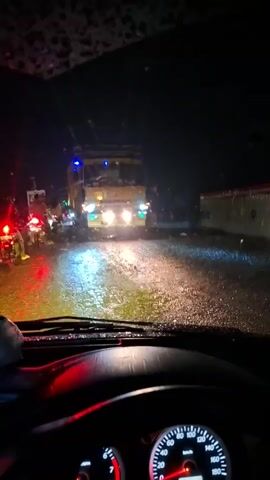 The video is filmed from inside a vehicle at night. The windshield is wet, with raindrops visible. The headlights of the vehicle illuminate a flooded road, with water splashing as vehicles move. A large yellow truck is directly ahead, its headlights bright. To the left, several motorcycles are visible, with their red taillights reflecting on the wet road. The dashboard of the car is in the foreground, with a speedometer showing a speed of approximately 18 kilometers per hour and a fuel gauge. The scene depicts a traffic situation on a rainy night, likely a traffic jam.