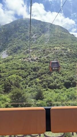 The video captures a scenic view from inside a gondola lift cabin, ascending or descending a lush green mountain in Čavori, Montenegro, on a bright, sunny day. The camera is positioned looking out through the large windows of the cabin, showing the expansive mountainous landscape covered in dense trees and vegetation. Above, a clear blue sky with fluffy white clouds adds to the picturesque setting.

In the foreground, the orange padded seats and black frame of the gondola cabin are visible, providing a sense of being high above the ground. Several thick cables stretch across the frame, guiding the gondola lift system. Another gondola cabin, dark orange and black, is seen passing by on the parallel cable, with the text "PEARL COASTER" visible on its side. The camera pans slightly to the right, revealing more of the rolling hills and a glimpse of the distant horizon. The overall impression is one of a serene and elevated journey through a beautiful natural environment.
