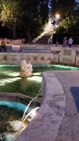 The video pans across a fountain at dusk. The fountain has multiple spouts and a central sculpture. The water is illuminated by underwater lights, creating a greenish glow. In the background, there are stairs leading up to a pathway lined with trees and lights. A few people are visible sitting on benches and walking on the pathway.