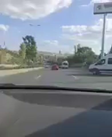 This video shows a first-person perspective from inside a vehicle, driving on a road. The scene is a city with buildings in the background, suggesting an urban or suburban setting. Trees and foliage line the side of the road, indicating a potentially mild or warm climate. The sky is blue with scattered clouds, suggesting daytime, likely mid-morning or afternoon given the lighting.

Several vehicles are visible on the road, including a white van directly in front of the camera's view, and a red car further ahead. The presence of multiple vehicles suggests moderate traffic. The camera appears to be looking forward and slightly to the right, capturing the road and the surrounding environment.

The overall impression is a mundane, everyday drive through a city. There are no people clearly visible within the frame, other than those likely inside the vehicles. The video does not reveal any specific events or activities beyond the act of driving. The location context provided places this scene in Mamak, Turkey. There is no visible text in the video.