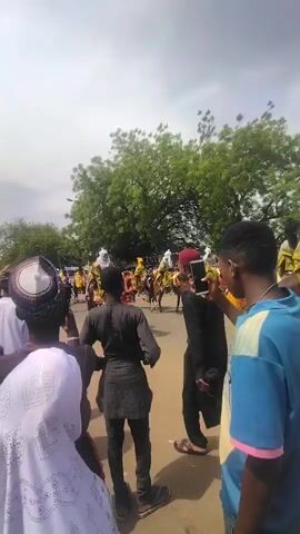 The video shows a large group of people gathered on a street. Some are on horseback, wearing colorful traditional attire. Many are on foot, watching the procession. Some are taking pictures with their phones. The sky is overcast, and there are trees in the background. The atmosphere seems festive and celebratory.