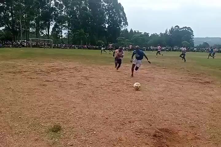 Here is a detailed description of the video:

The video shows a soccer game taking place on a dry, dirt field with patches of grass. There is a crowd of spectators along the sidelines, including people sitting and standing. In the foreground, a soccer player wearing a blue jersey, white shorts, and red socks dribbles a soccer ball. He is closely followed by another player wearing a pink jersey and black shorts. The player in blue collides with the player in pink, causing him to lose control of the ball. After the collision, the player in the blue jersey appears to watch the other player run towards the soccer ball.  Behind the players and the spectators, there are trees and a structure that resembles a building.