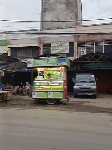 The video shows a street scene with several small shops and food stalls. The shops are mostly two-story buildings with open fronts displaying goods. Several food carts are visible, some with illuminated signs and displays of food items. People are seen walking and sitting around the stalls, and a few motorcycles are parked. The street is paved, and there are overhead power lines and a satellite dish visible. The overall atmosphere appears to be a local market or commercial area.