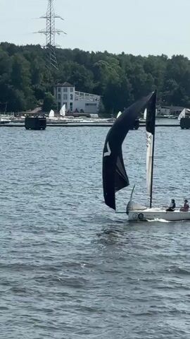 This is a video of sailboats on a body of water. In the foreground, a white sailboat with a blue sail is visible, with several people on board. In the middle ground, a smaller white sailboat with a large black and white sail is seen, also with people on board. In the background, trees and buildings line the shore, and a large electrical tower is prominent. Several other sailboats are visible in the distance. The water is choppy, indicating wind. The overall scene appears to be a sailing event or race on a sunny day.