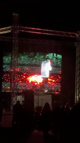 The video captures an outdoor night event in Balgari, Bulgaria, where an audience watches a large LED screen. The screen displays a performance featuring a person in a white garment interacting with vibrant red and orange glowing elements, resembling fire or embers, on a green, foliage-like background. The screen is supported by a robust metal truss structure. Silhouetted figures of the audience are visible in the foreground, indicating they are watching the display. A distinct musical beat is audible in the background.
