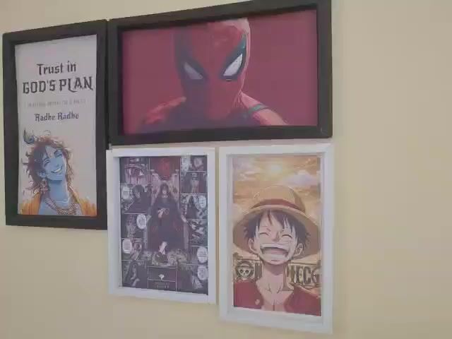 A wall displays four framed pictures. On the left, a large black-framed picture features a quote "Trust in GOD'S PLAN EVERYTHING HAPPENS FOR A REASON Radhe Radhe" above an illustration of a blue-skinned Krishna with a peacock feather in his hair. To the right of that, a horizontal black-framed picture shows a close-up of Spider-Man's masked face against a red background. Below the Spider-Man picture, two smaller white-framed pictures are arranged side-by-side. The left one is a collage of various anime characters, with a prominent central image of a character with dark hair and red eyes. The right picture shows a smiling anime character with a straw hat and a red shirt, with a bright sun and clouds in the background. The wall is a light, neutral color.