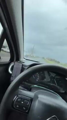 The video shows a view from inside a car driving on a paved road. The dashboard is black and includes a steering wheel with control buttons and a dashboard with an electronic speedometer. A phone is mounted to the left of the steering wheel, displaying a navigation app. The road ahead is straight, flanked by fields and some sparse trees. The sky is overcast and gray. The car is moving at a moderate speed. The video appears to be shot by the driver.
