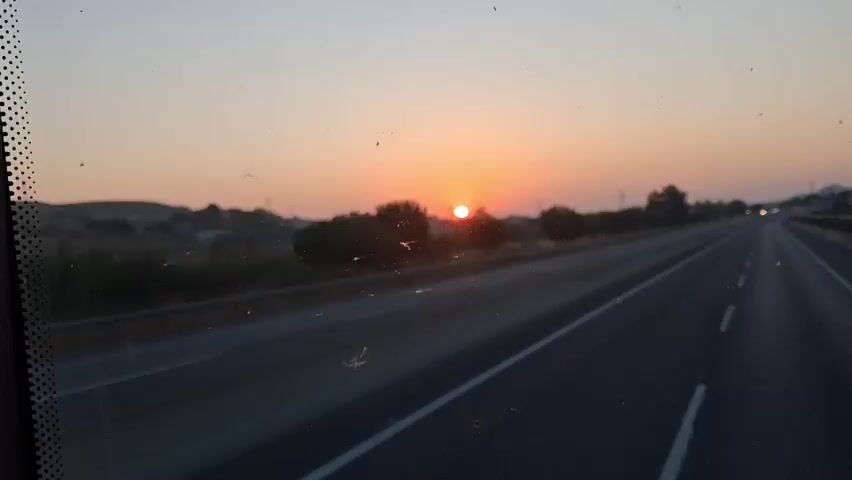 The video shows a view from inside a vehicle traveling on a highway at sunrise. The sun is a bright orange disc low on the horizon, casting a warm glow across the sky. The highway has multiple lanes, and a guardrail separates the road from the adjacent landscape. Trees and some buildings are visible on the left side of the road. The sky transitions from a pale orange near the sun to a lighter shade of pink and gray towards the top of the frame. The windshield appears to have some smudges or specks on it. The overall impression is one of peaceful travel during the early morning hours.