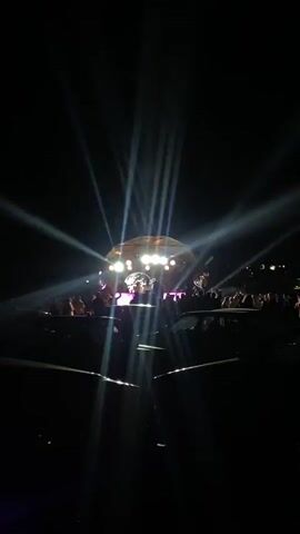 The video shows a night concert with bright stage lights illuminating the performers and a crowd of people. The stage appears to be a dome-shaped structure with multiple lights. The crowd is visible in silhouette as they stand in front of the stage. The camera is positioned behind the crowd, looking towards the stage.