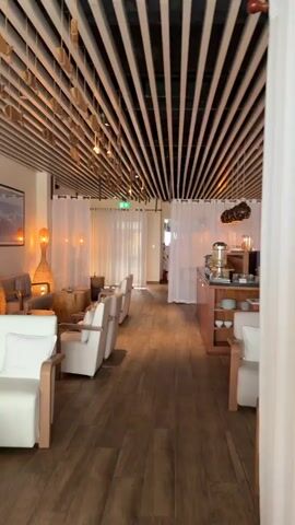 The video pans across a modern, well-lit interior space that appears to be a lounge or waiting area. The room features a warm and inviting ambiance, with natural wood tones and soft lighting. The ceiling is adorned with horizontal wooden slats, creating a visually appealing pattern. Several plush white armchairs with wooden frames are arranged throughout the space, some with small wooden tables in front of them. White sheer curtains hang from the walls, adding a touch of elegance and softness. On the walls, framed artwork depicting tropical scenes, including overwater bungalows and serene ocean vistas, enhances the tranquil atmosphere. Potted plants, such as a palm, are strategically placed to add a touch of greenery. Wicker lamps with warm orange light bulbs are visible, contributing to the cozy mood. The floor is made of dark wood planks, which complements the overall aesthetic. In the background, a counter area with a beverage dispenser and shelves stocked with items suggests a service area, possibly for drinks or refreshments. The space is clean, organized, and designed for relaxation and comfort.