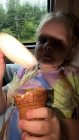 A young girl with blonde hair in pigtails sits in a vehicle, holding an ice cream cone. She has a drip of ice cream on her chin. The window behind her shows a blurry green landscape. A hand enters the frame from the left, offering a spoon with ice cream. The girl looks at the spoon. The video is shaky and dimly lit.