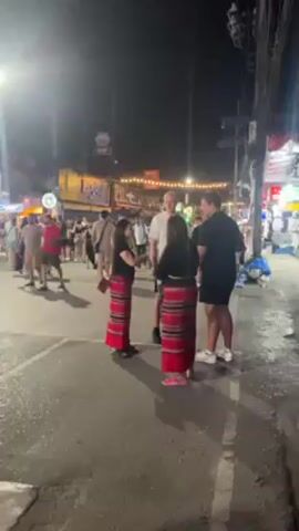 The video depicts an outdoor nighttime scene on a bustling street. The primary focus is a group of four individuals engaged in conversation. Two women, wearing matching outfits of black tops and long red skirts with striped patterns, stand facing two men. One of the men is wearing a white t-shirt and the other is wearing a black outfit. The street is crowded with people moving in both directions, and various shops and stalls are visible in the background, illuminated with bright lights and signs. Overhead, strings of decorative lights add to the vibrancy of the scene. The street surface is dark and appears to be asphalt. Overall, the scene suggests a lively and busy urban setting at night.