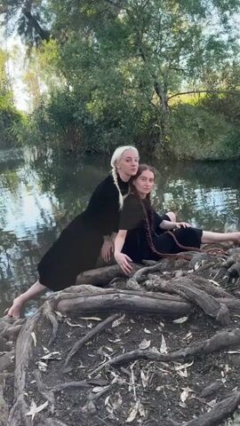 Two young women wearing long black dresses are posing by a body of water. The woman on the left has blonde hair styled in a braid and is leaning on the woman on the right, who has brown hair also in a braid. They are sitting on a pile of exposed tree roots on the bank of the water. The water is calm and reflects the surrounding trees and foliage. The environment appears to be a natural setting with trees and vegetation visible in the background. The lighting suggests it is daytime.