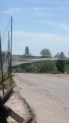 The video shows a rural scene in daylight, possibly in a conflict zone. Anti-tank obstacles, also known as Czech hedgehogs, are visible along the side of a partially paved road. Overhead, a net-like structure is suspended between poles, presumably for protective purposes. The netting appears to be white or light-colored. Across the road, dense vegetation lines the other side. Behind the Czech hedgehogs, there are glimpses of buildings, including a weathered wooden structure and a house painted in light blue. Power lines are also visible overhead. The scene suggests a location experiencing tension and security measures.