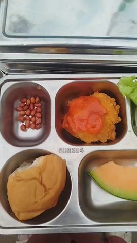 The video shows a close-up overhead view of a partitioned stainless steel lunch tray filled with food. The tray is divided into five compartments. In the top left compartment, there is a pile of peanuts. In the compartment to its right, there is a piece of fried food topped with a red sauce. To the right of that, in the top right compartment, there is a piece of lettuce and a slice of white onion. In the bottom left compartment, there is a bread roll. In the bottom right compartment, there is a slice of cantaloupe melon. The words "SUS304" are visible embossed in the metal of the tray, below the fried food compartment. The background is slightly blurred and appears to be an outdoor setting, possibly a table.