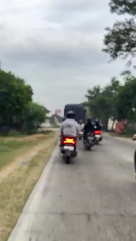 This is a first-person perspective, vertical video of a motorcycle procession on a two-lane road. The camera person is riding a red scooter, followed by several other scooters, and a large truck is visible ahead. On the left side of the road, there is a dirt shoulder with some bushes and trees, and a few buildings are visible in the distance. On the right side of the road, there are more trees and some buildings. The sky is overcast. The scooters are moving at a steady pace, and the traffic appears to be light. There are at least five other scooters visible in the video.