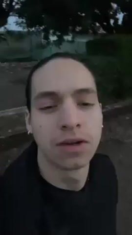 A person with their hair pulled back is recording a selfie video outside. The person is wearing a black jacket and has small hoop earrings. They are standing on a pathway near some greenery and what appears to be a building in the background. The sky is visible and appears to be dusk.