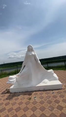 A white statue of a woman in a flowing robe is positioned on a patterned tiled ground. The statue is centrally located in the frame, with a body of water and a treeline visible in the background. The sky above is blue with scattered clouds. The camera pans slightly to the right, providing a wider view of the statue and its surroundings.