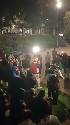 Here's a detailed description of the video:

The video shows a group of approximately 25 people gathered at night in what appears to be a park or public outdoor space. The scene is illuminated by overhead streetlights and at least two portable spotlights that are very bright, casting strong shadows. Most individuals are dressed casually in jackets, hoodies, and hats. A few people are clapping, while others hold up their phones as if recording. In the center of the crowd, one person wearing a red vest and grey pants appears to be the focus of attention, likely speaking or performing. There seems to be a small area covered with artificial turf on the ground. Behind the group, there are trees, a path, and a bench visible, suggesting it is an urban park environment. The overall mood seems celebratory or focused on an event taking place in the center of the group.