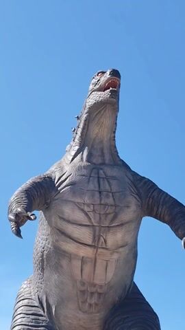 The video shows a static, low-angle shot of a large, gray Godzilla statue against a clear blue sky. The statue is depicted with its head raised and mouth open, as if roaring. Red color is visible inside the mouth and in the eyes of the statue. Its arms are outstretched.