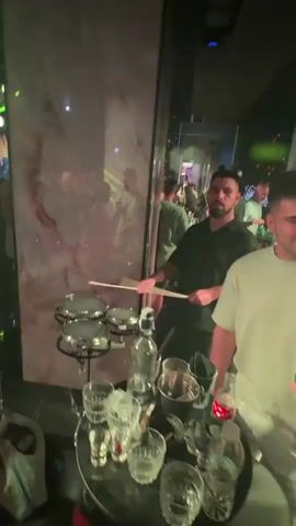 The video shows a dimly lit indoor environment, possibly a club or bar, with colorful, illuminated panels. In the foreground, a man with dark hair and a beard is playing a small drum set with drumsticks. He is wearing a dark shirt. Next to the drum set are several bottles, including what appears to be a bottle of Finland vodka, and various glasses. In the background, other people are visible, some standing and others blurred. The lighting shifts throughout the video, with areas of white light, and later a predominantly blue light. One shot shows a person handing the drummer something, possibly a drink or another item. Another shot shows the drummer with his head bowed towards the drum set, and the blue lighting creates a silhouette effect. The overall atmosphere appears to be one of a social gathering or nightlife.