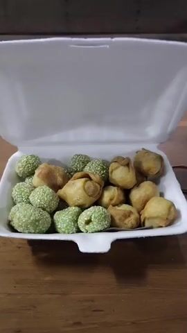 A white styrofoam take-out container is filled with a variety of fried snacks. The container is open, with the lid propped up. The snacks are arranged in the bottom of the container and include small, round, green pastries coated in sesame seeds, as well as a few golden-brown, fried dumplings. The container sits on a wooden surface, likely a table. The lighting suggests an indoor setting.