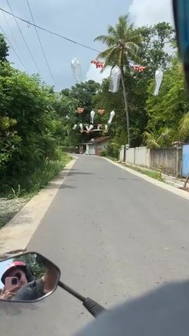 A motorcycle POV shot shows a paved road winding through a lush, green area, likely tropical, with trees and overgrown vegetation. The road is decorated with Buddhist flags and white paper lanterns strung overhead, creating a festive atmosphere. A single building is visible on the right side of the road. In the motorcycle’s left side mirror, a person wearing a pink helmet is visible holding a smartphone. The sky is partly cloudy. The camera shakes consistently, likely from the motion of the motorcycle.