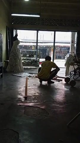 A person is crouching on the floor in front of a large window. The person is wearing a yellow t-shirt and dark pants. They appear to be working with some wooden planks on the floor. To the left of the person, a white dress is displayed on a mannequin. Through the window, a street scene is visible with shops and passing vehicles. The room is dimly lit, with light filtering in from the window and a light fixture overhead. The floor is concrete and appears worn in places.