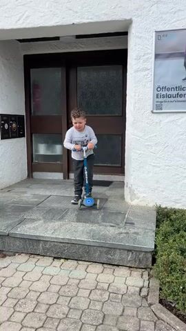 A young boy with curly brown hair, wearing a gray long-sleeved shirt and dark pants, stands on a blue three-wheeled scooter at the top of a short flight of stone steps in front of a building. The building has a white facade and dark double doors for an entrance. A poster is visible on the wall to the right of the doors. The boy, looking forward and smiling, propels himself and the scooter down the two steps onto a paved walkway below. He continues moving forward on the scooter as the video ends.