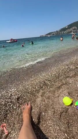 The video depicts a sunny day at a pebble beach, likely in Bečići, Montenegro. The foreground is dominated by a person's bare foot and a collection of colorful children's beach toys, including a pink truck, a yellow ball, a pink shovel, and a blue shovel. The sand is a mix of small pebbles and some darker granules.

In the middle ground, the clear, turquoise water of the Adriatic Sea laps gently against the shore. Several people are visible in the water, some swimming and others wading. A red boat with a white canopy is anchored near the shore.

The background reveals a bustling beach scene. A line of beach umbrellas and loungers suggests a popular tourist area. Further in the distance, a town with white buildings climbs up a green, mountainous hillside under a bright blue sky dotted with a few white clouds. The overall atmosphere is one of leisure and enjoyment, typical of a summer vacation. The time of day appears to be midday, given the strong sunlight and shadows. There is no visible text in the video.