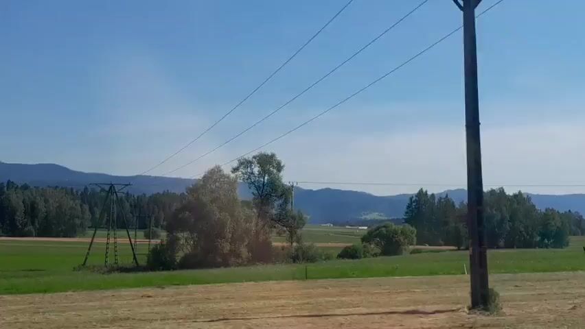 The video shows a pastoral landscape with green fields, trees, and hills under a clear blue sky. Power lines stretch across the frame. A tractor is visible in the distance, appearing to be in a field. The overall impression is of a rural, agricultural setting.