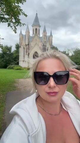A woman with blonde hair wearing large black sunglasses and a white hoodie is taking a selfie in front of a small, ornate church with a pointed steeple. The church appears to be old and weathered, with a white facade and a wooden door. It is surrounded by a green lawn and trees. The sky is overcast. The woman's face is partially obscured by her sunglasses and the camera angle.