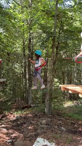 Two young girls, secured by harnesses and wearing helmets, navigate an aerial ropes course in a forest. The girl on the right, in a blue helmet, is crossing a suspended wooden plank bridge, holding onto ropes for balance. The girl on the left, in a white helmet, is standing on a circular wooden platform attached to a tree, reaching out to the other girl. Lush green trees surround them under a bright sky.