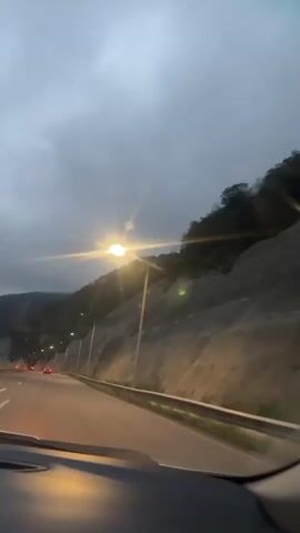 A dash cam view from a moving vehicle on a highway. The sky is cloudy and overcast. The sun is visible as a bright light, creating lens flares. A mountainous terrain with rocky slopes and trees is visible on the right side of the road. Streetlights line the highway. Several vehicles with red taillights are driving ahead on the road. The interior of the car is visible in the foreground, including the dashboard and windshield. The video appears to be shot in the early morning or late evening due to the dim lighting and the presence of streetlights.