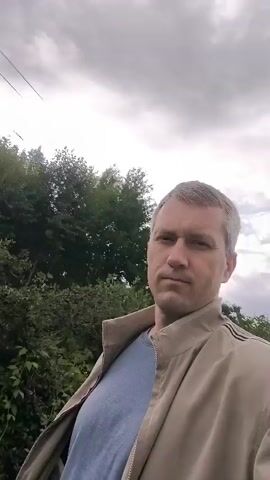 A man is seen outdoors in front of a slightly cloudy sky. He is wearing a beige jacket over a blue t-shirt. In the background, there are trees, grass, and a house. The man is looking directly at the camera. The video appears to be a selfie.