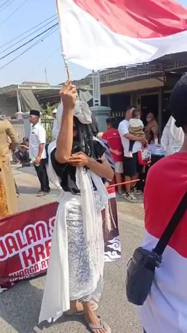 A person dressed in a white sheet draped over their head and body, with long dark hair visible, is holding a red and white flag and a stick. The person is wearing patterned leggings and flip-flops. They are standing on a street, possibly during a parade or festival, as there are other people in the background, some of whom are also dressed in costumes or holding flags. A banner with red lettering is visible in the foreground. The environment appears to be outdoors, with buildings and a clear sky.