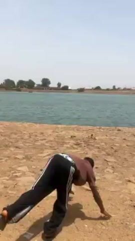 A young man is performing a handstand on a rocky shoreline in front of a lake. He's wearing a brown t-shirt and black track pants with white stripes. He briefly loses his balance, returns to standing, then takes a few steps forward. The lake is calm and stretches across the background, with sparse trees and buildings visible on the far shore under a clear sky.