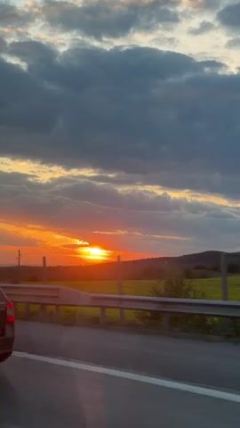 The video shows a sunset viewed from a car on a highway. The sun is low on the horizon, partially obscured by clouds. Fields are visible to the side of the road, and a highway guardrail is in the foreground. A black Skoda Octavia is briefly visible to the left. A large billboard is visible to the left, with text in Bulgarian. The car appears to be moving.