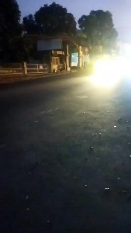 The video begins at dusk or dawn on an asphalt road. The camera is positioned low to the ground, facing forward. In the foreground, the road surface is dark and shows some debris. In the background, there is a building with several structures or windows. A bright light, likely from a vehicle's headlights, illuminates a portion of the road and the building. The sky is a dim gray. The scene appears to be an outdoor, possibly rural or suburban, environment. There are no visible people in the frame.