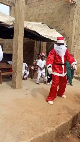 A man in a Santa Claus costume dances in an outdoor setting surrounded by children. He is wearing a red suit, white gloves and boots, and a Santa hat, and his face is covered with a white beard and sunglasses. Other people are standing and sitting nearby, including a man in white traditional garb who is filming and laughing. The setting appears to be a courtyard with unfinished brick buildings around. Some children dance along to the music.