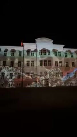 At night, a projection shines onto the facade of a light-colored, multi-story building. The projection displays an abstract image with red, blue, white, and earth-tone colors, appearing to move and shift on the surface of the building. A flag is visible on the roof of the building, and the rest of the environment is dark.
