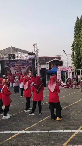 This is a vertical, eye-level shot of an outdoor event. In the foreground, four people are standing facing away from the camera. The person on the left is wearing a blue shirt and black pants, with a black hijab. The three people to their right are wearing red shirts and black pants, with red hijabs. In the midground, a group of people, also mostly wearing red and black attire, are gathered in front of a stage. The stage has a banner that reads "SEMARAK KEMERDEKAAN REPUBLIK INDONESIA" and features images of Indonesian historical figures and flags. There are two large speaker towers on either side of the stage. To the left of the stage is a blue tent, and to the right are several tents and more people. The background shows buildings and tall, slender trees. The ground is paved with interlocking bricks and has white lines painted on it, suggesting it might be a sports court or field. The lighting suggests it is late afternoon or early evening.