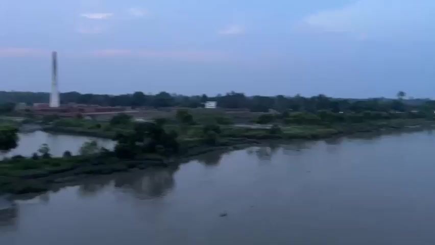 The video shows a wide, muddy river with a calm surface. The riverbanks are lined with green vegetation and trees. In the distance, there are some buildings and structures, suggesting a settlement or village along the river. The sky is a pale blue, indicating it might be early morning or late evening. The overall scene is peaceful and natural.