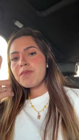 The video features a woman sitting in a car. She has long brown hair, fair skin, and is wearing a white t-shirt, pearl earrings, and a yellow necklace with a turtle pendant. She is making gestures with her hands, starting by pointing her index finger upward, then moving her hand to rest on her forehead, and finally making a peace sign. She smiles and moves her finger again. The car's interior is visible, including the ceiling, rear seats, and part of the dashboard. There is also background music playing.