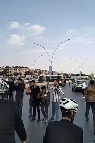 The video shows a group of people on bicycles gathered on a street, seemingly in a protest or demonstration. Many of them are wearing helmets. Some people are holding up their phones, apparently taking pictures or videos. Some individuals are wearing black clothing with green accents. A Syrian flag is briefly visible. The backdrop features buildings and streetlights. A vehicle is visible in the background. The overall impression is of a political gathering or demonstration.