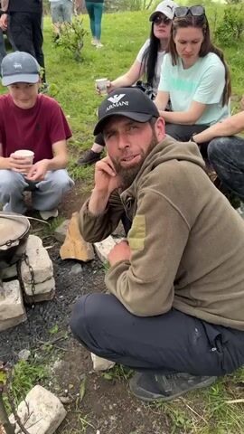 This is a vertical video of a group of people gathered outdoors. In the foreground, a man wearing a black baseball cap and a khaki-colored hooded jacket is crouched down, looking at the camera. He has a beard and is holding his hand to his chin. Next to him, to his left, a younger person wearing a maroon t-shirt and a blue baseball cap is also crouched down, holding a paper cup. A large pot sits on a makeshift stove made of bricks and metal chains in front of them. Behind the man in the foreground, several other people are visible, some sitting and some standing. They appear to be having a picnic or a gathering. A white off-road vehicle is parked in the background. The environment is grassy and appears to be a rural or natural setting with hills in the distance. The sky is overcast.