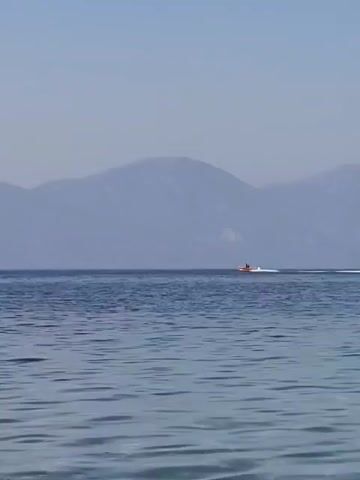 A small orange and white boat with two people onboard travels across calm blue water. In the background, a hazy mountain range is visible under a clear sky. The boat is moving from left to right, leaving a white wake behind it.