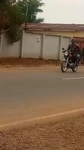 A person in a pink shawl walks away from the camera on a dirt shoulder next to a road. A motorcycle passes by on the road behind them. The person's face is not visible. A second person, a boy in a light-colored shirt and dark shorts, walks toward the camera on the same shoulder. He is carrying a bag over his shoulder. The road appears to be paved with a white line marking the center. Buildings and trees are visible in the background.