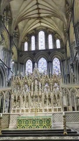 The video shows the interior of a large, ornate cathedral. The camera pans across the altar area, focusing on a highly decorated reredos with numerous sculptures and architectural details. Behind the reredos, stained-glass windows are visible, illuminated by natural light. The walls and ceiling feature Gothic architecture with intricate ribbed vaults and pointed arches. In the foreground, there is an altar cloth with a green and gold pattern. Candles are placed on stands to the right and left of the altar. To the left of the altar, a large floral arrangement is visible, along with several wooden chairs. The overall impression is one of grandeur and historical significance, with a focus on religious art and architecture.