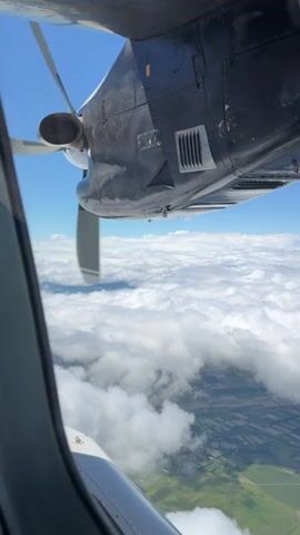 This is a first-person perspective video taken from an airplane window. The camera is positioned to look out of the window and also capture a portion of the airplane's wing and engine. A propeller is visible on the left side of the frame, appearing to be in motion. The airplane is flying above a layer of white clouds, and below the clouds, a landscape of green fields and some agricultural patterns can be seen. The sky is clear and blue. The overall scene depicts an aerial view during daytime flight.